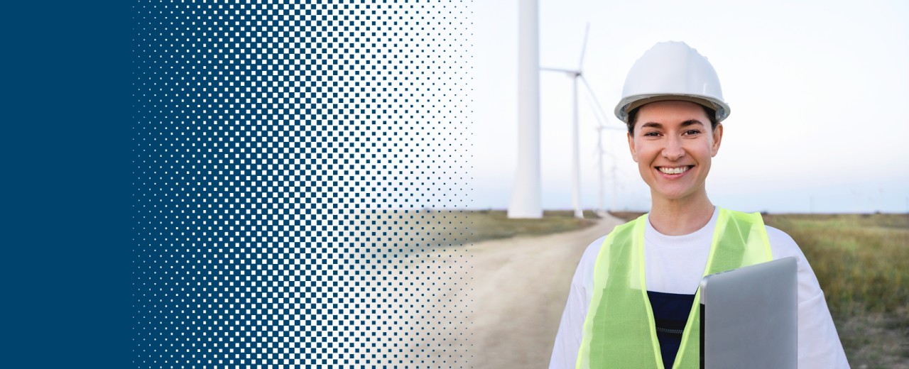 A woman wearing protective clothing and a hard hat stands in front of a wind farm with a laptop